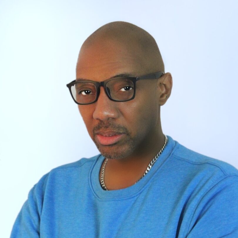 Alphonso King Jr folds his arms and looks seriously at the camera. He wears black rimmed glasses, a blue sweatshirt and a silver chain necklace.