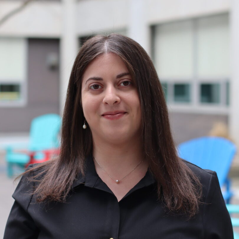 Anna Kozak stands outside and smiles at the camera, wearing a black button up shirt and simple pearl jewelry.