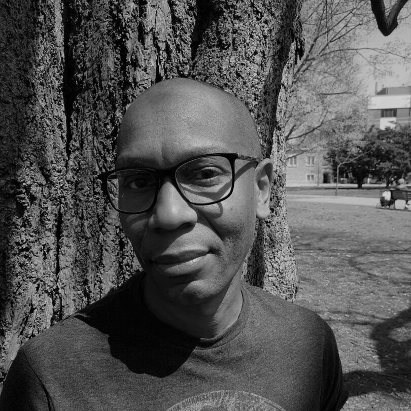 In a black and white photo, Brian stands in front of a tree and smiles slightly at the camera while wearing square rimmed glasses and a tshirt.