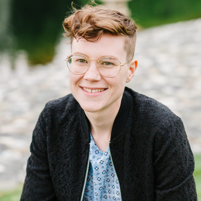 A young adult white non-binary person smiles at the camera. They have short curly brown hair and are wearing gold-rimmed glasses, a light blue patterned shirt, and a lace-textured black jacket.