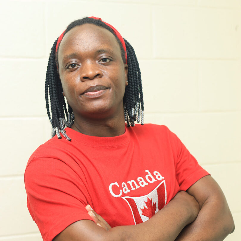 With arms folded, Christopher smiles slightly at the camera while wearing a bright red Canada tshirt with the Canadian flag on it and her hair in braids accessorized with clear beads.