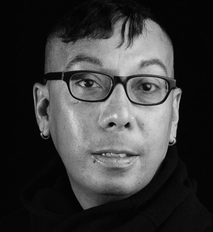 A black and white close up shot of Christopher Smith in a black turtleneck and black rimmed glasses, smiling slightly at the camera.