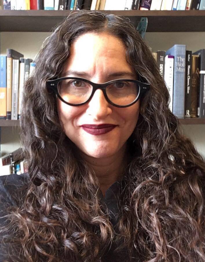 A headshot of Dana Seitler smiling slightly at the camera. She is a white woman with long dark brown curly hair and she wears black thick-rimmed glasses and matte burgundy lipstick. In the background are several shelves of books.
