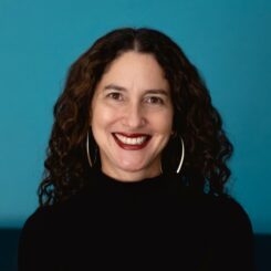 Dana Seitler smiles at the camera against a deep teal background. She wears large silver earrings and a brick coloured lipstick.