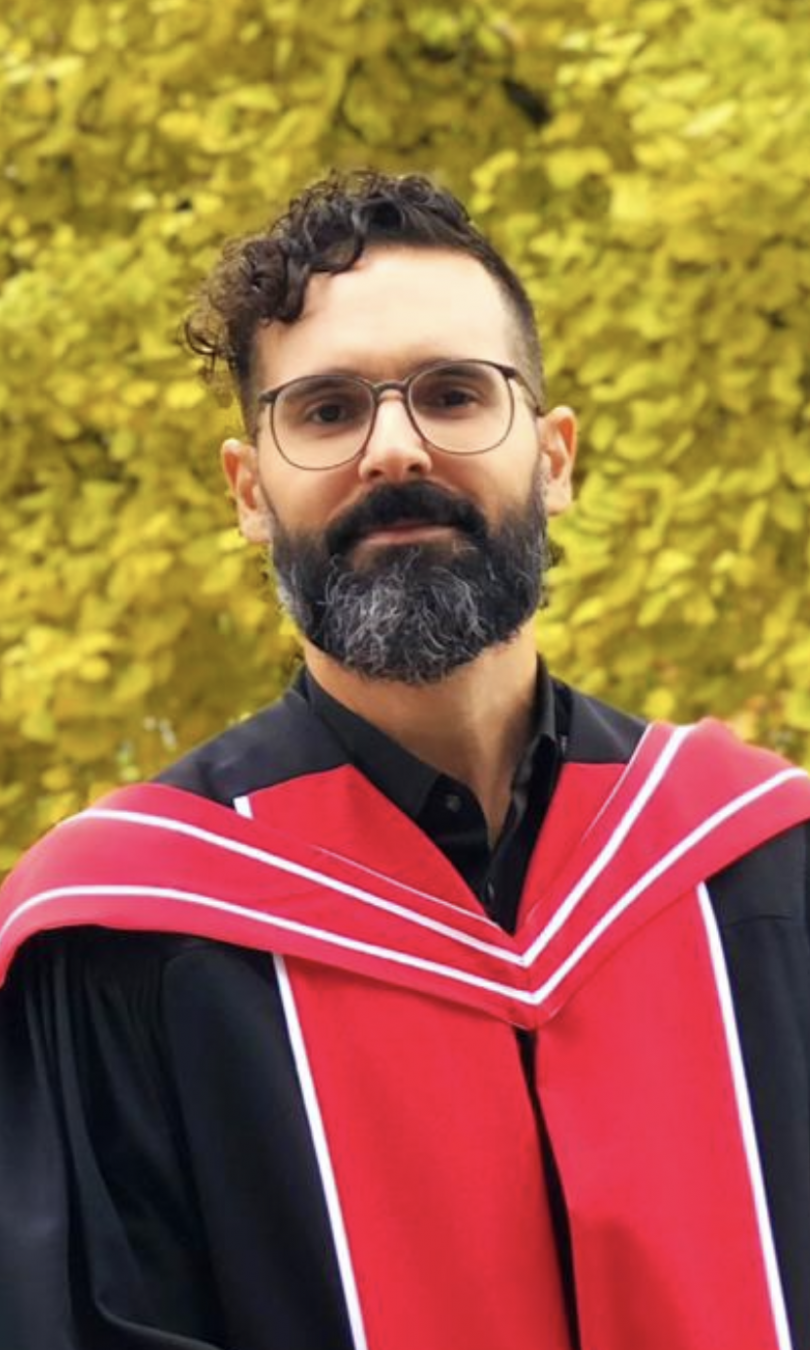Wearing a red and black academic regalia, David Pereira stands in front of a gingko tree with yellow leaves. He wears round horn-rimmed glasses and has a full beard and short, curly dark hair.