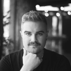 In a black and white photo with blurred lights in the background, Elliott smiles directly at the camera and poses thoughtfully with their hand by their chin.