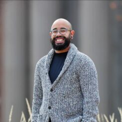 Qui Alexander smiles widely while standing outdoors, wearing a black turtleneck under a grey cardigan and blue square rimmed glasses.