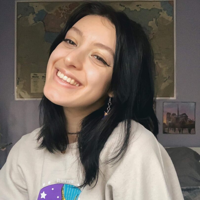 A person with straight dark hair is wearing a beige sweater with a blue and purple sun and moon design in the centre. She is tilting her head upwards to the left and smiling with teeth showing. There is a large world map taped to a lavender wall behind them in the background.