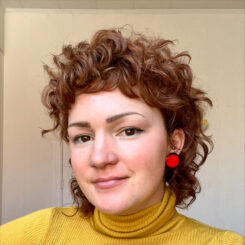 Hannah smiles slightly at the camera, wearing a mustard yellow turtleneck and a bright red round earring.
