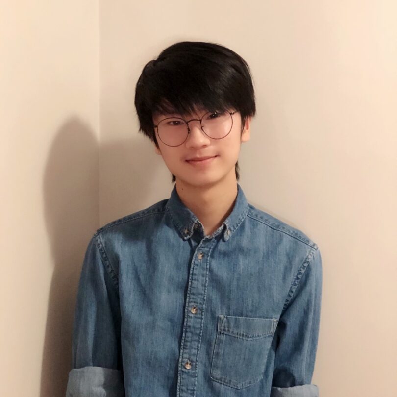 Henry Yang smiles at the camera, wearing a denim button up shirt and wire-rimmed glasses against a white wall.