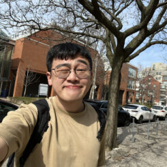 In direct sunlight, Ho Lam squints and smiles at the camera with one eye closed. He wears thin rimmed round glasses and a tan sweatshirt.