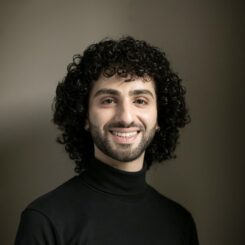 Jad smiles widely directly at the camera, wearing a black turtleneck.