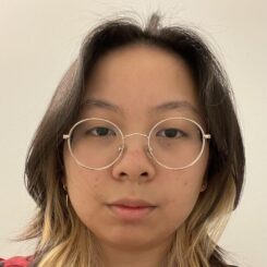 Janice looks pensively at the camera while wearing round gold-framed glasses.