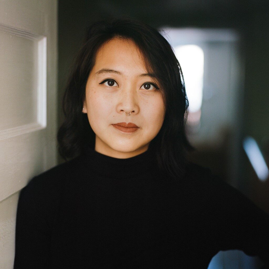 Jody smiles slightly at the camera while wearing black eyeliner, brick lipstick, and a black long sleeve shirt.