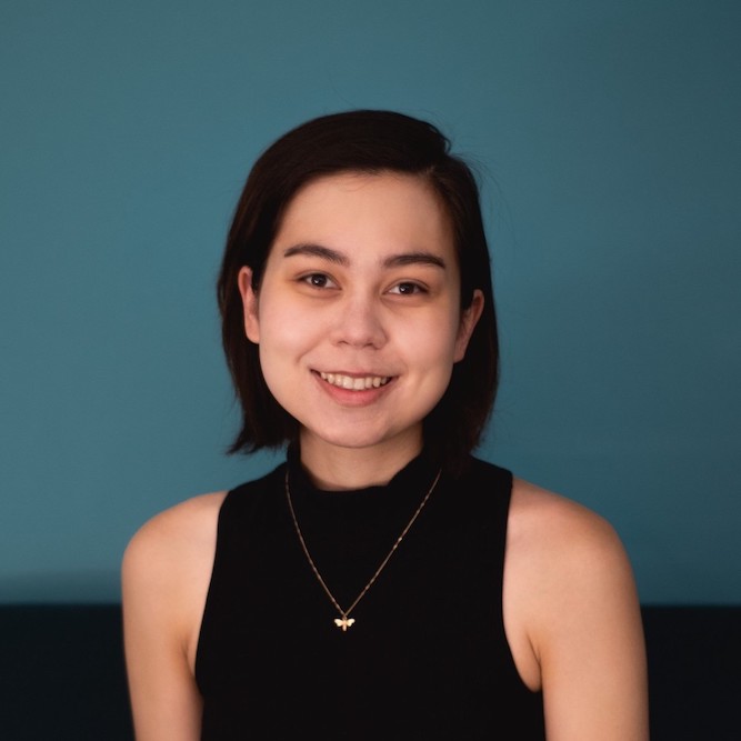 A headshot of Kanika, a biracial Asian queer person with short dark hair. They are wearing a black sleeveless sweater with a gold bee necklace. They are seated in front of a teal background.