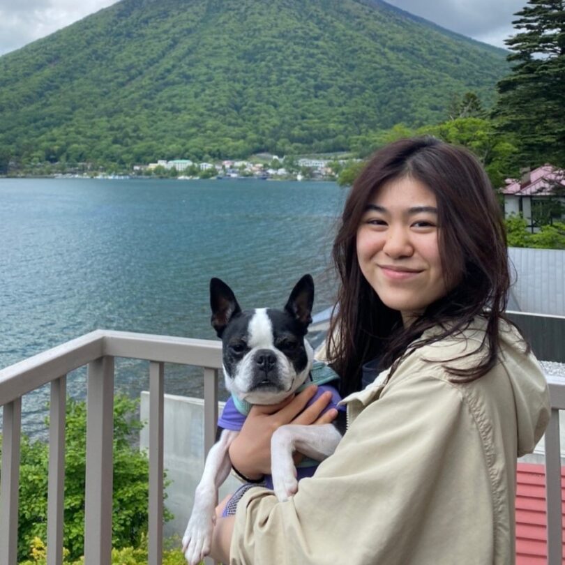 Luna Okazaki holds a small dog and smiles in front of a lake and a mountain in the background.