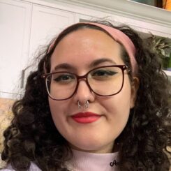 Olivia smiles at the camera, wearing black cat eye liner, red lipstick, and large framed glasses.