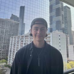 Ollie smiles at the camera in front of backdrop of skyscrapers, wearing a backwards baseball cap and a necklace with a sword pendant.