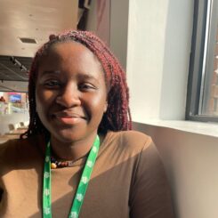Wearing a brown long sleeved shirt and green lanyard, Osarumen smiles at the camera with a window shining sunlight onto the side of the face.