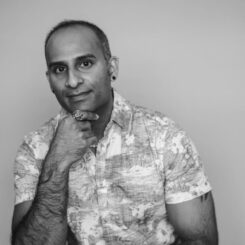 In a black and white photo, Jordache smiles at the camera while posing thoughtfully with his hand by his chin.