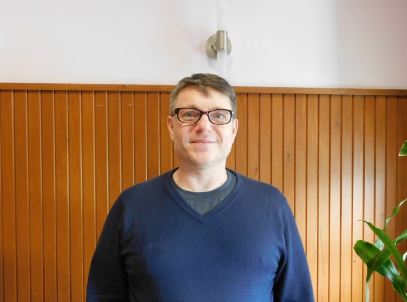 Scott Rayter smiles at the camera while wearing a blue v-neck sweater and brown rectangular glasses. His light brown hair is cut short and he stands a wood panelled background. 