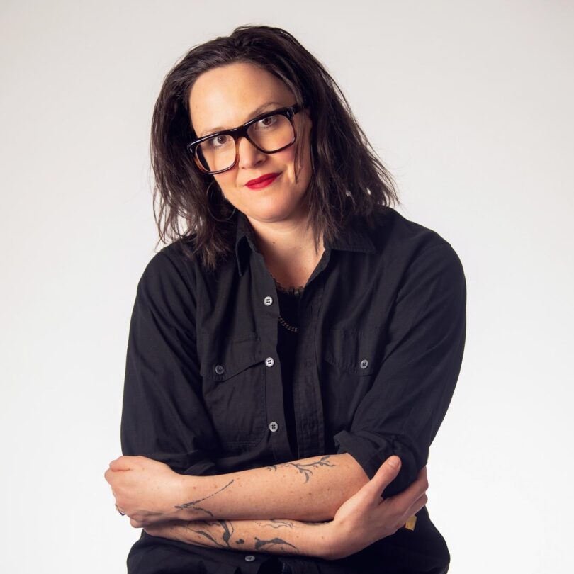 TL Cowan smiles at the camera with arms slightly crossed in a portrait shot. TL wears large square rimmed glasses, red lipstick, and a black button up shirt.
