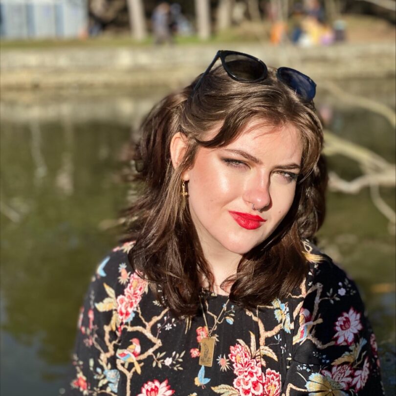 Photo of a person in a floral dress looking into the camera with a pond out of focus behind them.