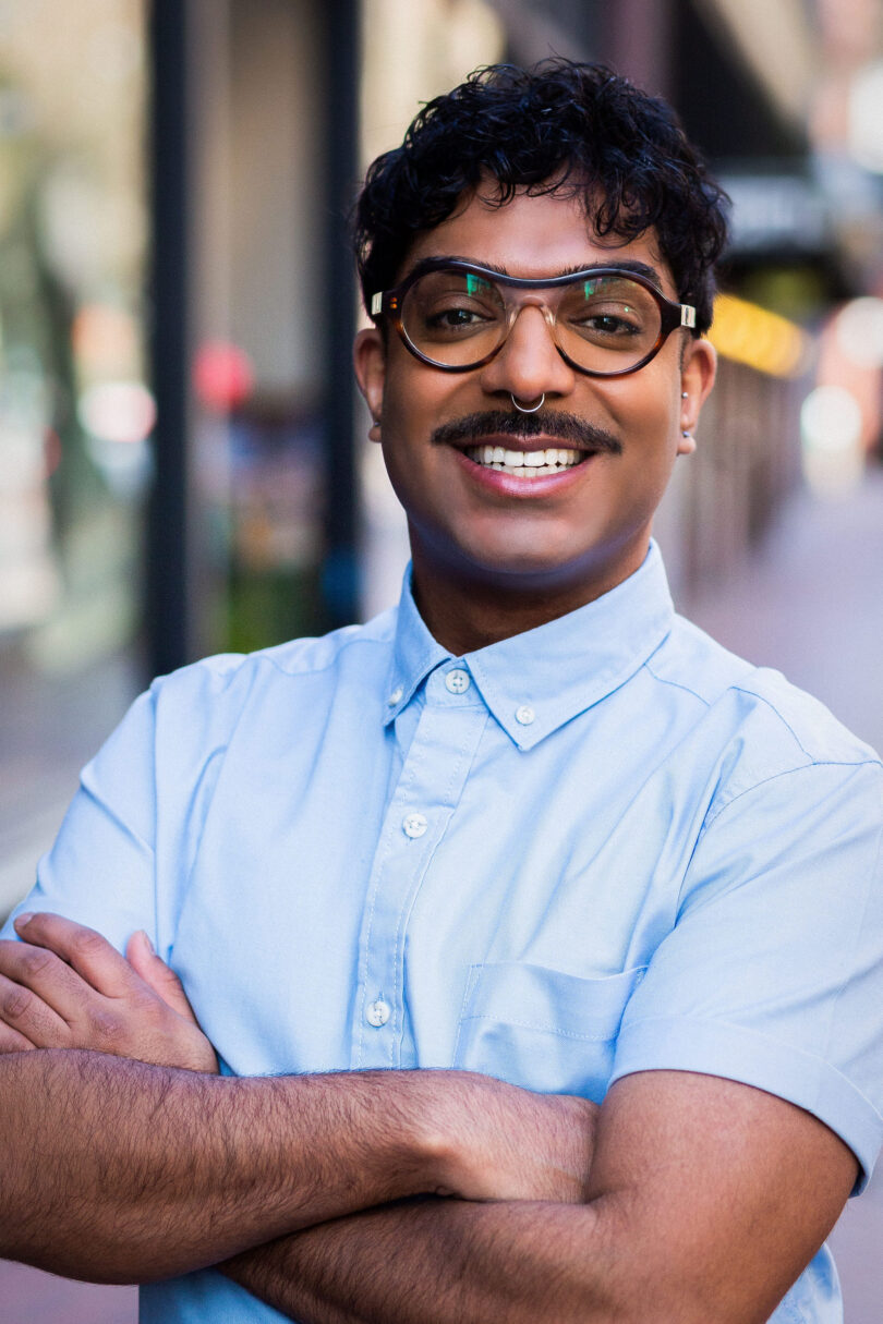 Portrait shot of Rahim Thawer smiles widely at the camera. He wears thick rimmed round glasses and a blue collared shirt. HIs arms are crossed.