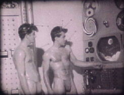Black and white photograph of two nearly nude men posing and examining a machine.