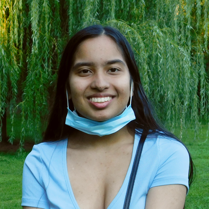 Anissa faces the camera and smiles widely, wearing a light blue mask around the chin and light blue shirt against a backdrop of a green park and willow tree.