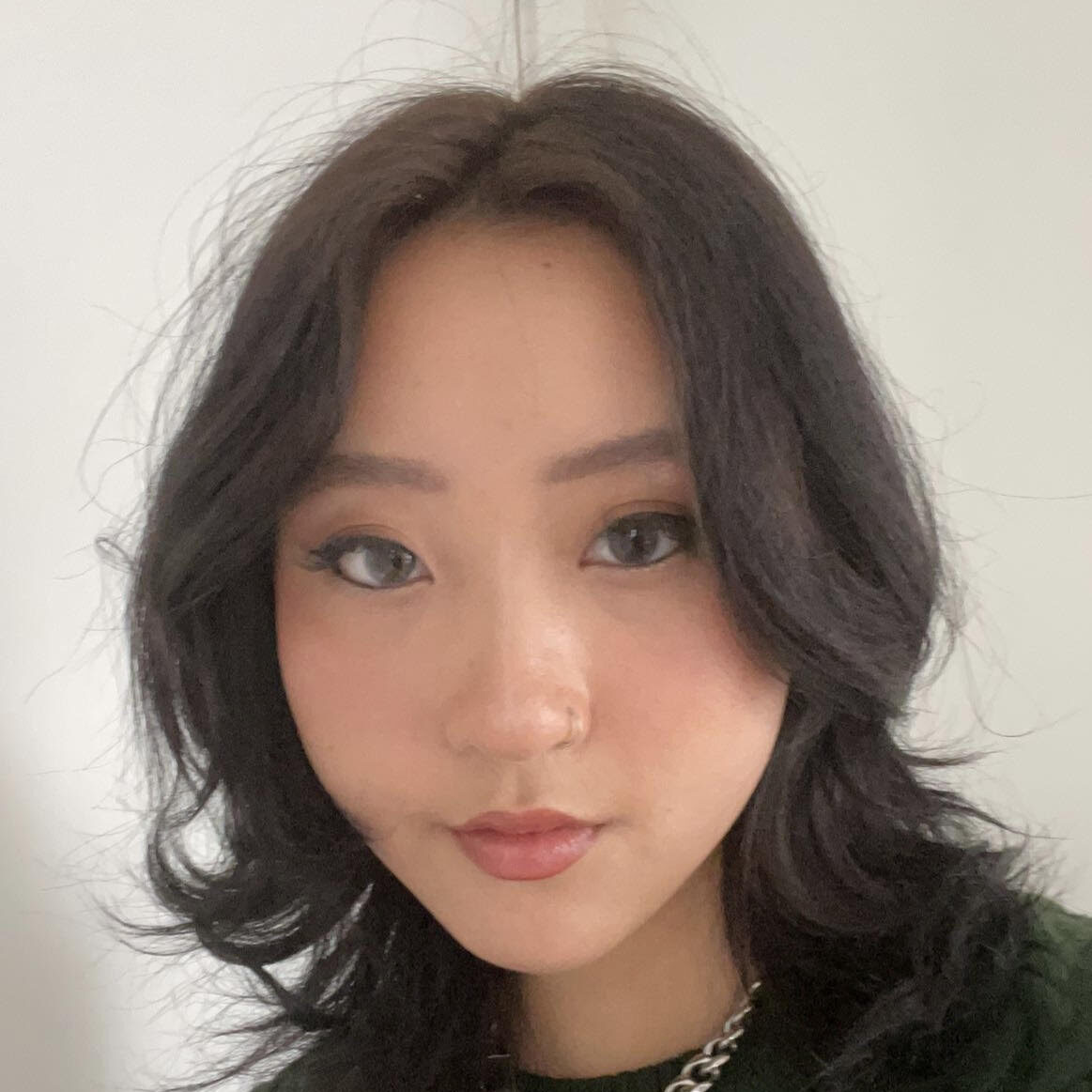 Carrie smiles slightly at the camera, wearing a thin silver chain necklace and deep green sweater. Carrie has shoulder length black hair and black eyeliner on.