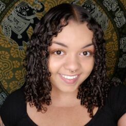 Chelle smiles widely at the camera. They have shoulder length curly hair and wear a black tshirt, with a tapestry in the background. 