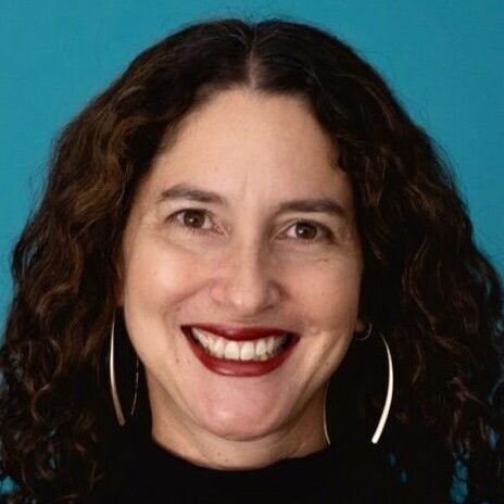 Dana Seitler smiles at the camera against a deep teal background. She wears large silver earrings and a brick coloured lipstick.