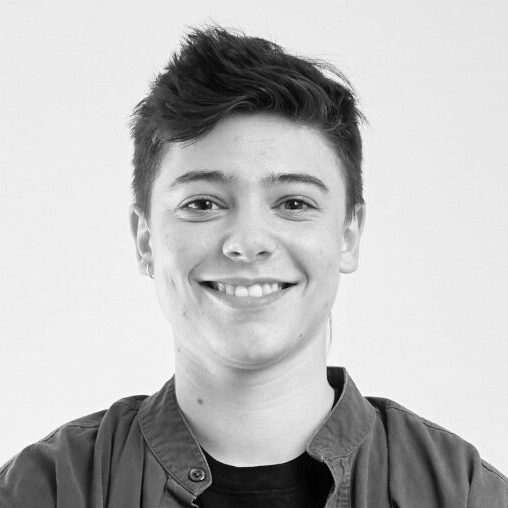 Black and white headshot of Elio Colavito from the chest up smiling in a t-shirt under an open button up shirt.