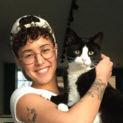 Holding a black and white cat, Maddie smiles at the camera while wearing a backwards baseball cap and round glasses. 