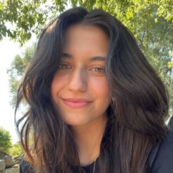 Riya Joshi smiles slightly at the camera against a sunny outdoor background. 